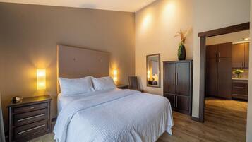 A hotel room with a large bed, wooden floor, a nightstand with a lamp, a wardrobe, and a mirror.