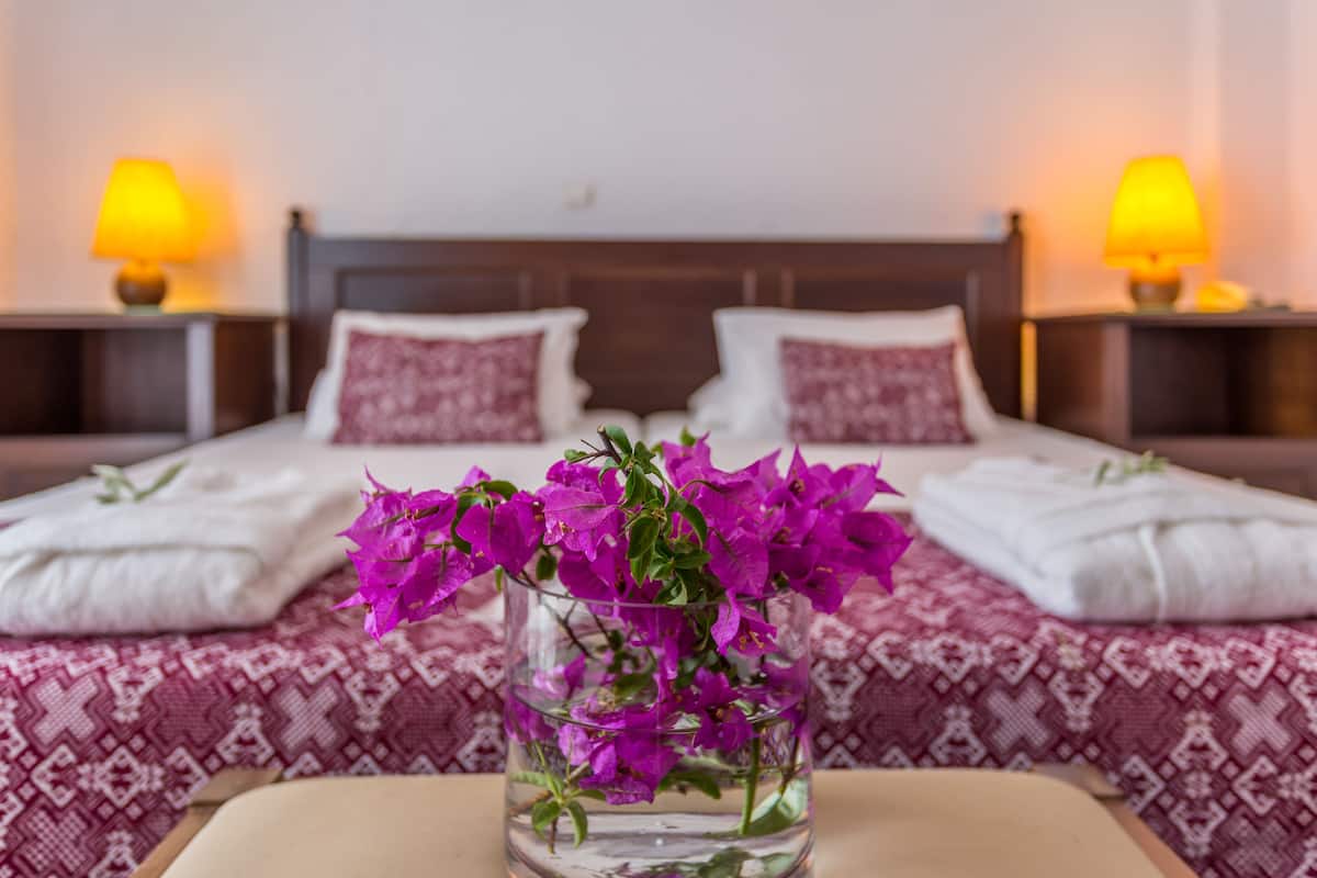 Una habitación de hotel con una cama, almohadas, una mesita de noche con lámpara y un florero con flores.
