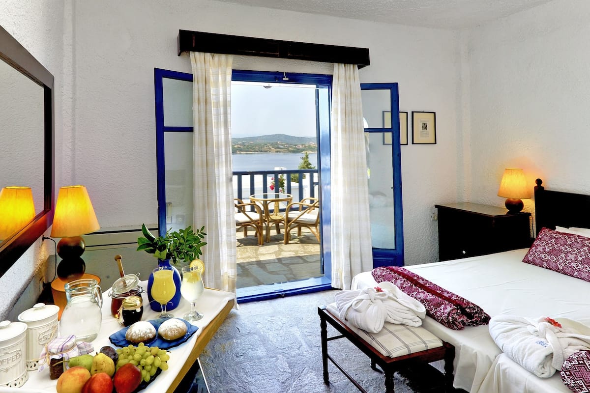 Habitación de hotel con cama, una mesa con frutas y bebidas, un balcón con mesa y sillas, y vista al mar.