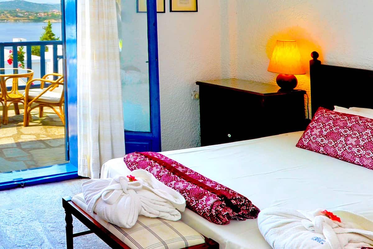 Una habitación de hotel con balcón, una cama con manta de diseño rojo y blanco, una mesita de noche con lámpara, un jarrón con flores y una bandeja de desayuno con pasteles y bebidas.