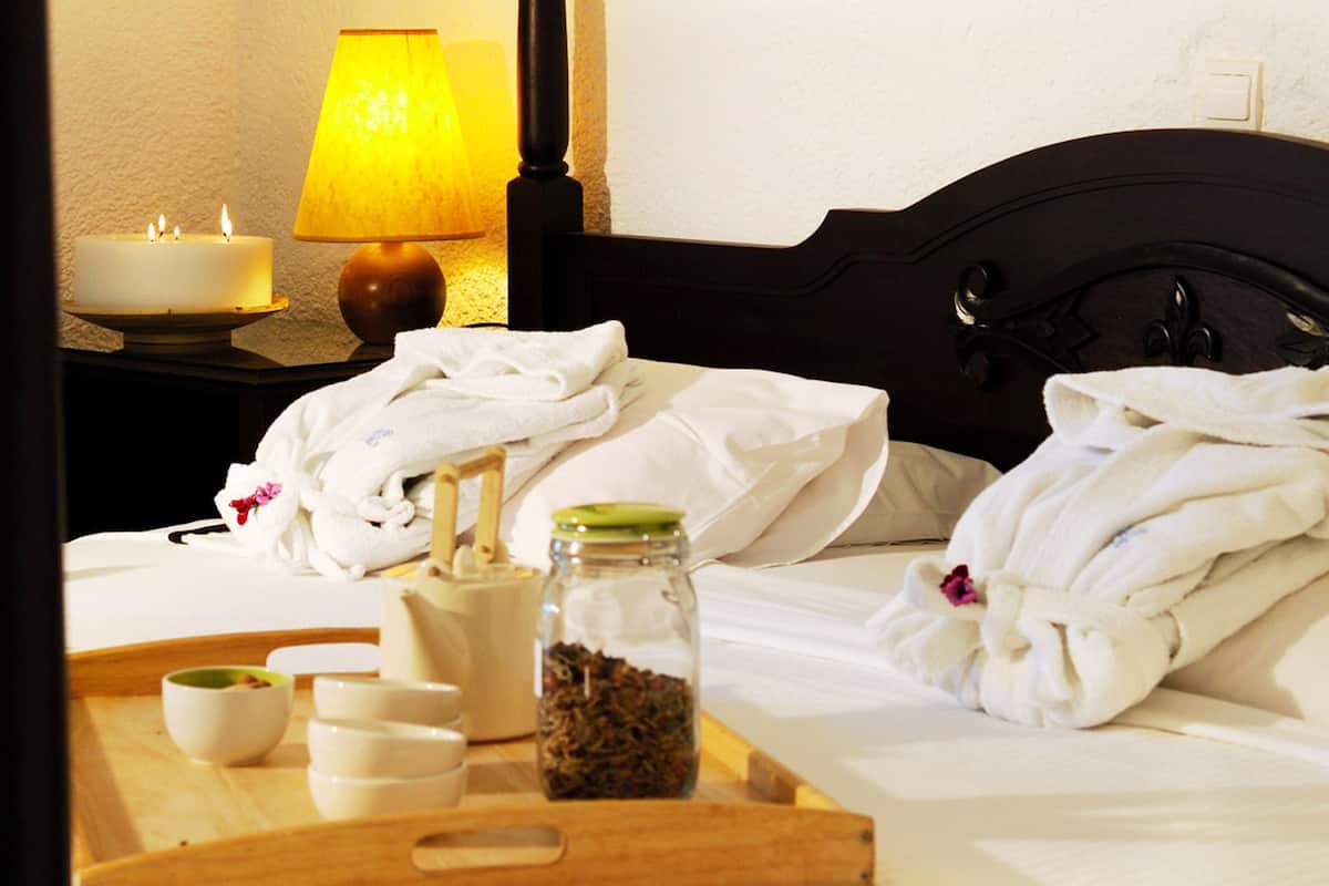 Una cama bien hecha con sábanas blancas, una lámpara de noche, una vela y una bandeja con artículos de tocador, incluyendo un tarro de té, una botella y recipientes pequeños.