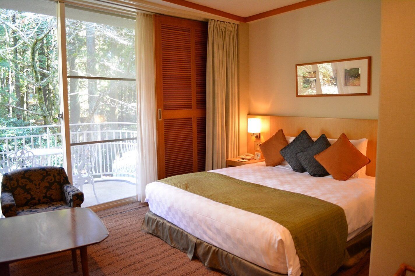 Hotelkamer met een groot bed, een bureau, een stoel, een balkon met uitzicht op bomen en een ingelijst schilderij aan de muur.