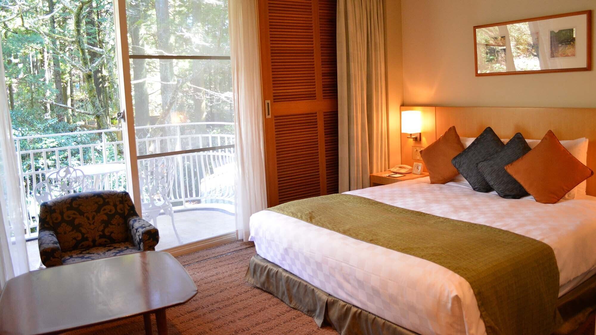 Ein Hotelzimmer mit Bett, Sessel, Tisch, einem Balkon mit Geländer und einem Vorhangfenster.