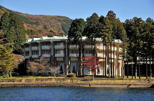 Hotel de vários andares com varandas, situado perto de um corpo d'água com montanhas ao fundo.