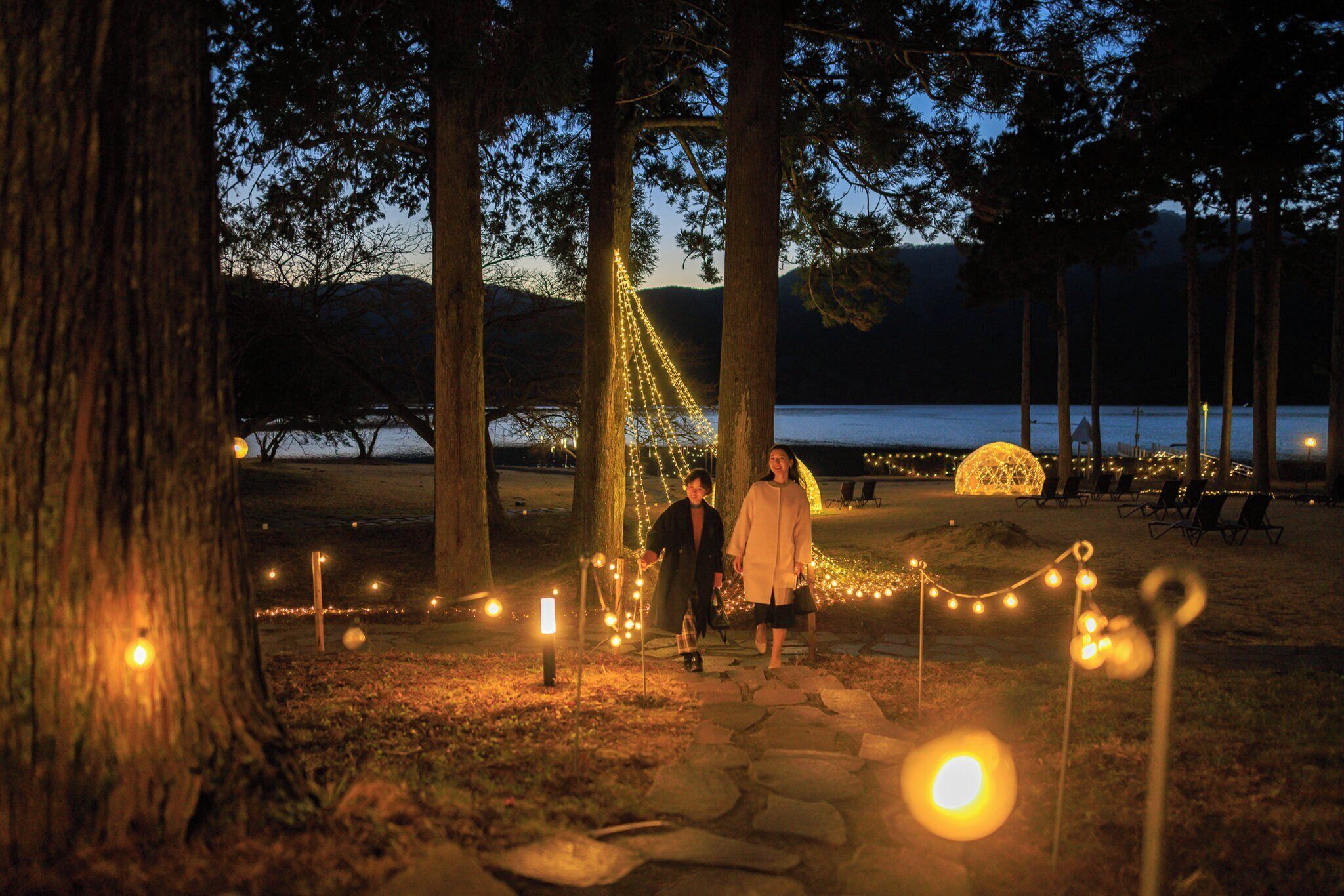 A pathway lined with trees and string lights leading to a gazebo by a lake at dusk.