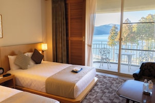 Quarto de hotel com cama, mesa de cabeceira, uma mesa com uma garrafa e vista para o mar e montanhas através de grandes janelas.