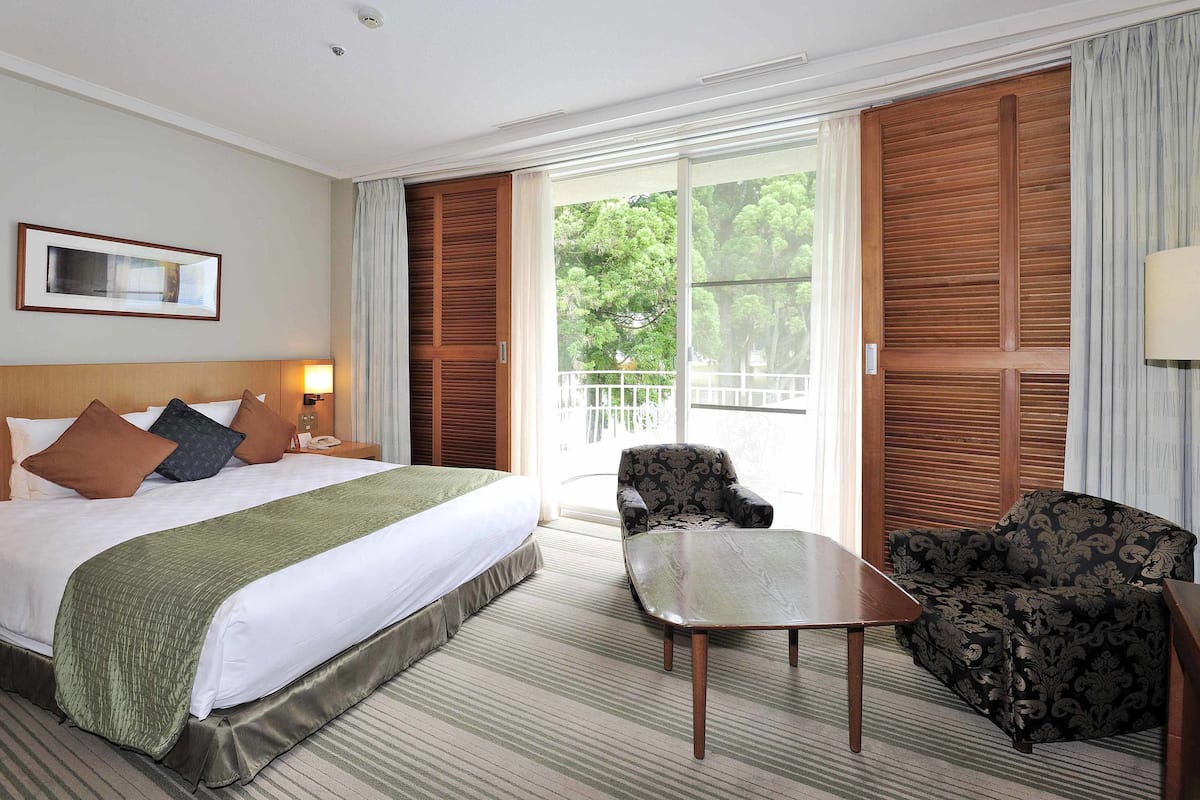 Habitación de hotel con cama, una silla, una mesa y un balcón con vistas a árboles.