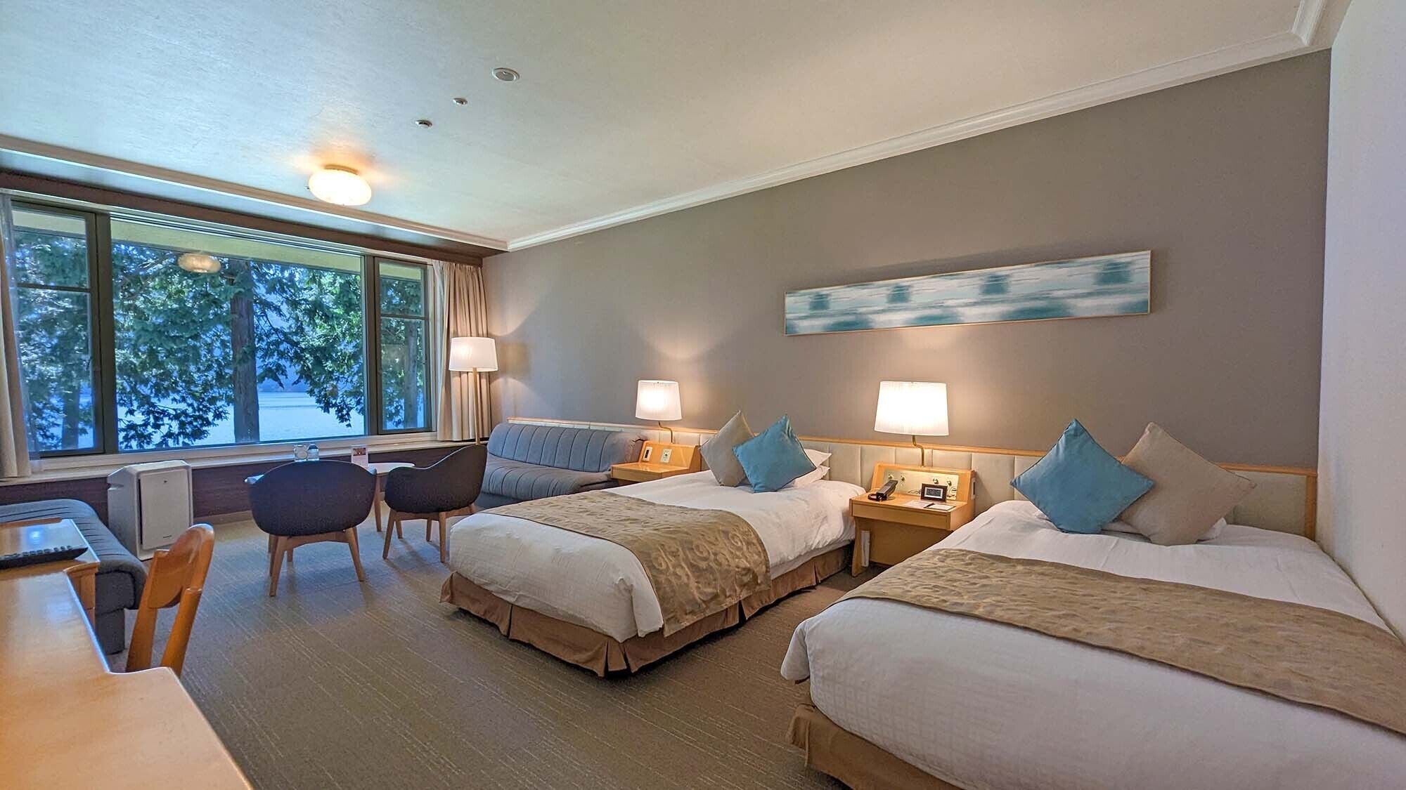 Ein Hotelzimmer mit zwei Betten, einem Schreibtisch, Stühlen und einem Fenster mit Blick auf Bäume.