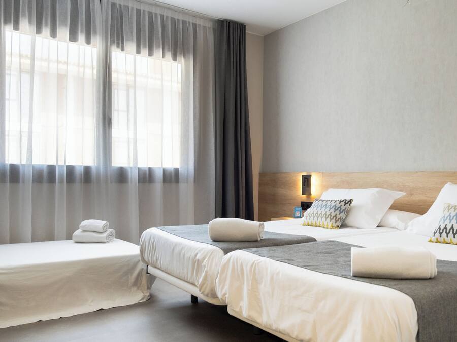 Habitación de hotel con dos camas, un ventanal con cortinas translúcidas, cabecera de madera y mesita de noche con lámpara.