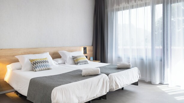 A modern hotel room with two beds, a wooden headboard, and sheer curtains.