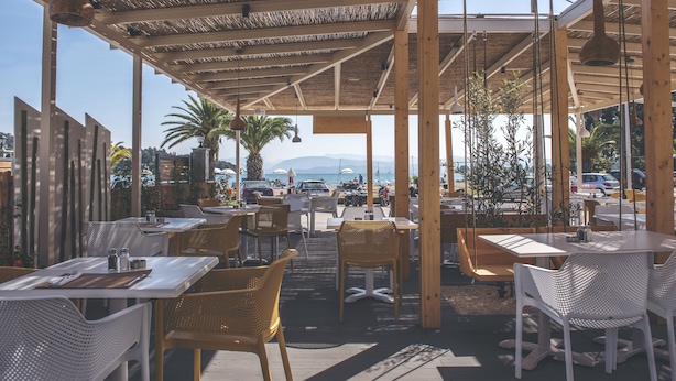 Ein Straßencafé mit Tischen und Stühlen, einem Reetdach und Blick auf Meer und Berge.
