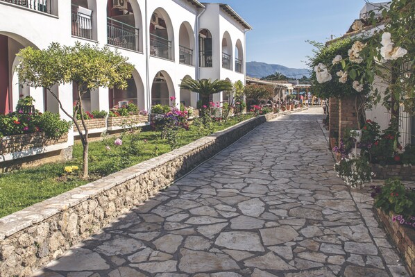 A stone-paved street with a white building featuring arched windows and balconies, surrounded by lush greenery and flowering plants.