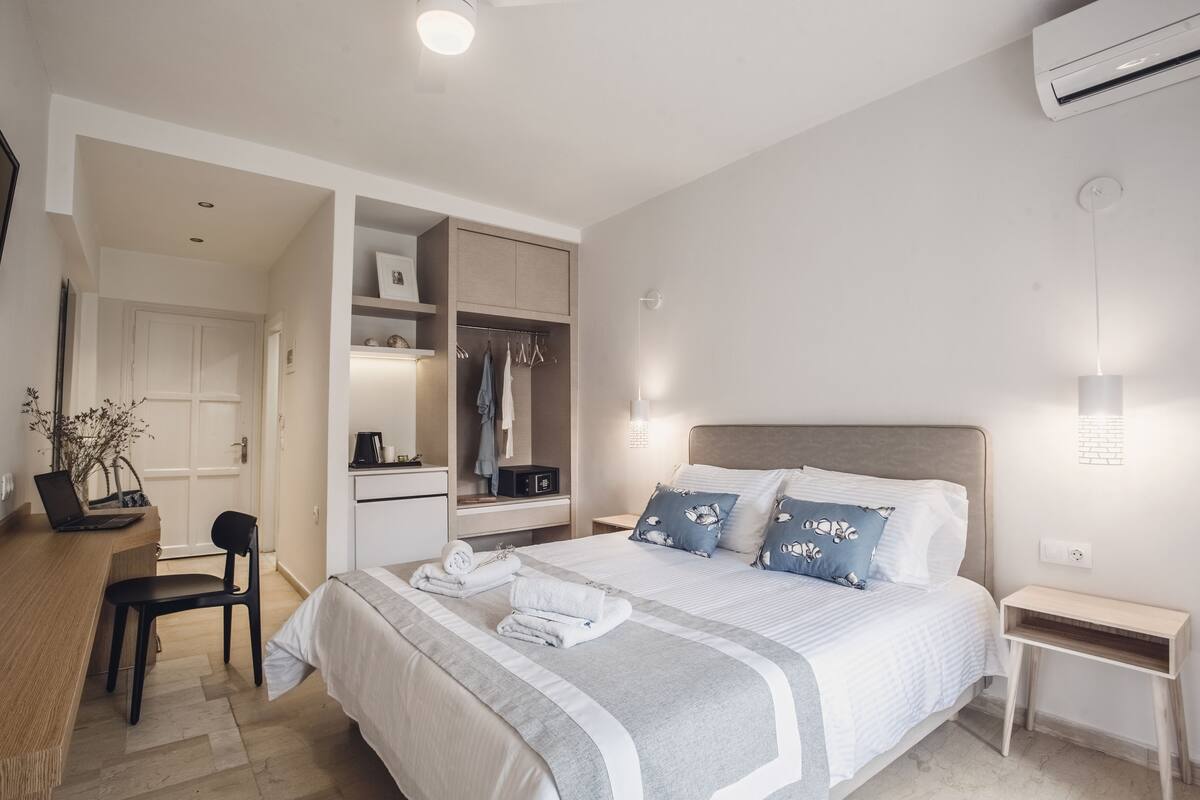 A modern hotel room with a neatly made bed, a desk with a laptop, a chair, and a wardrobe.
