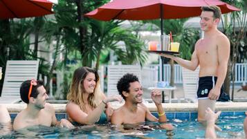 Une zone autour d’une piscine où des personnes nagent et se détendent à l’ombre de parasols rouges.