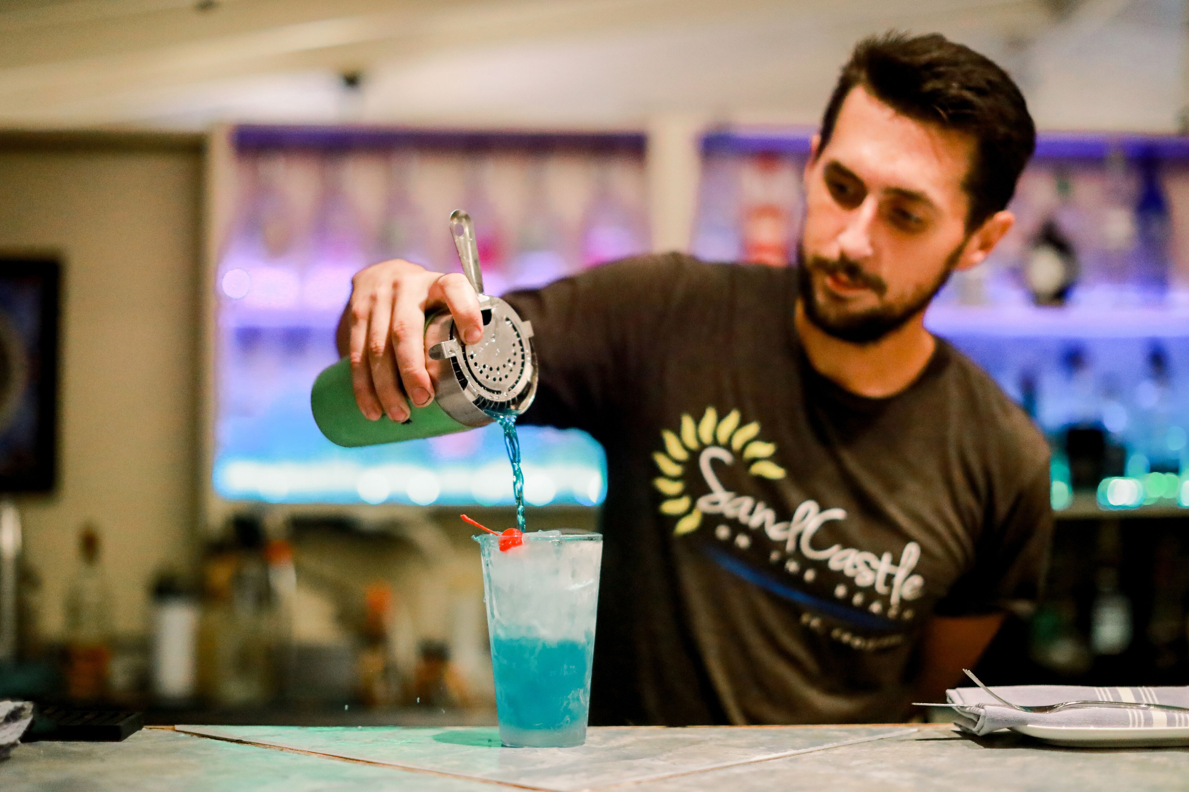 Ein Barkeeper gießt ein blaues Getränk in ein Glas mit Eiswürfeln und einer Kirsche.