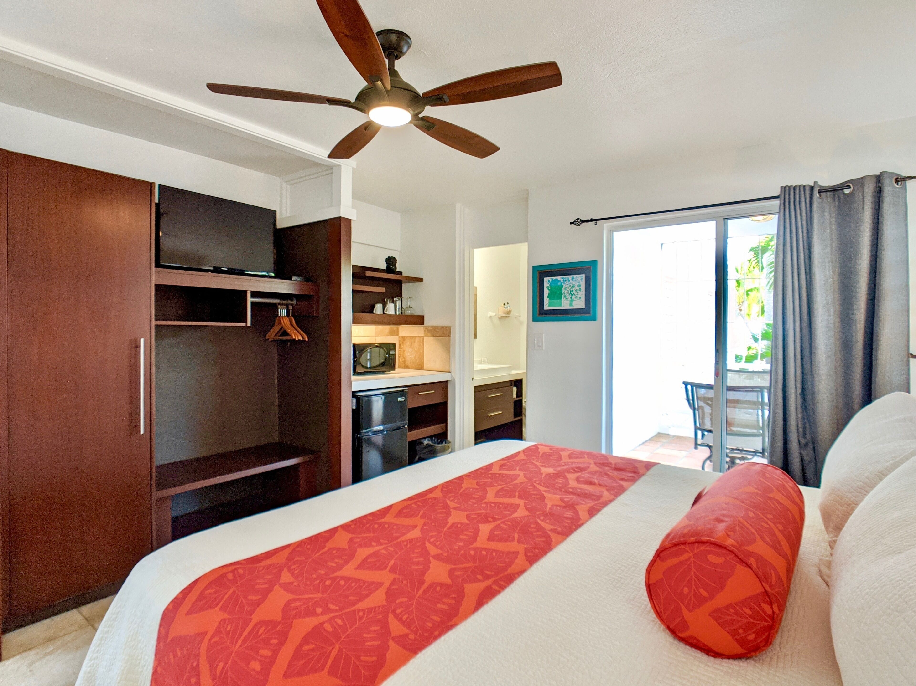 A bedroom with a bed, a ceiling fan, a wardrobe, a kitchenette, and a balcony with curtains.