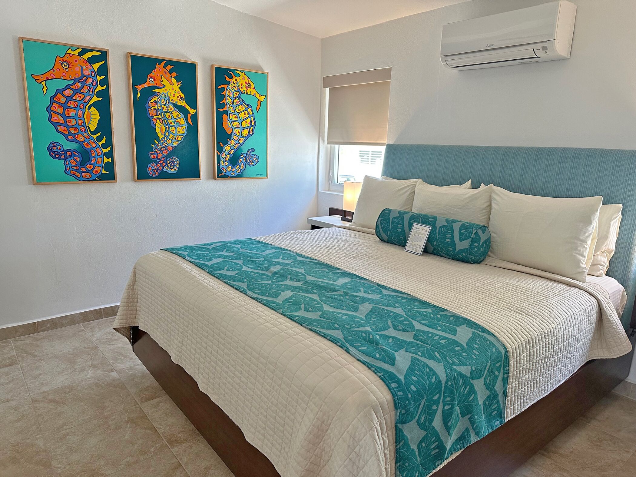 A hotel room with a bed, a nightstand, an air conditioner, and three colorful seahorse paintings on the wall.