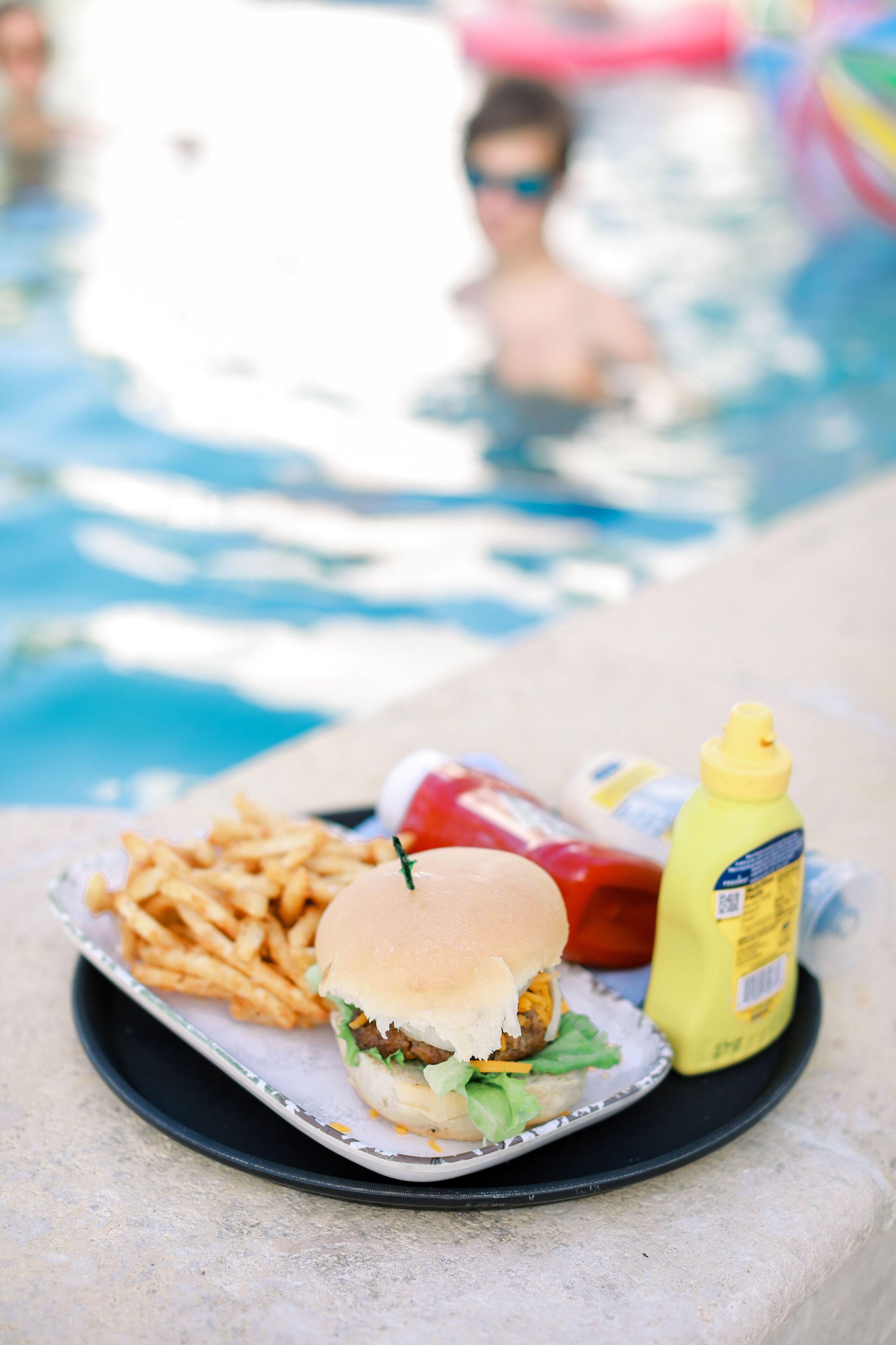 Comida que incluye una hamburguesa, papas fritas, kétchup y mostaza, sobre una bandeja cerca de una piscina.