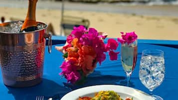 A table set with a plate of food, a glass of water, a bottle of champagne, and a flower arrangement on a beach with a blue umbrella.