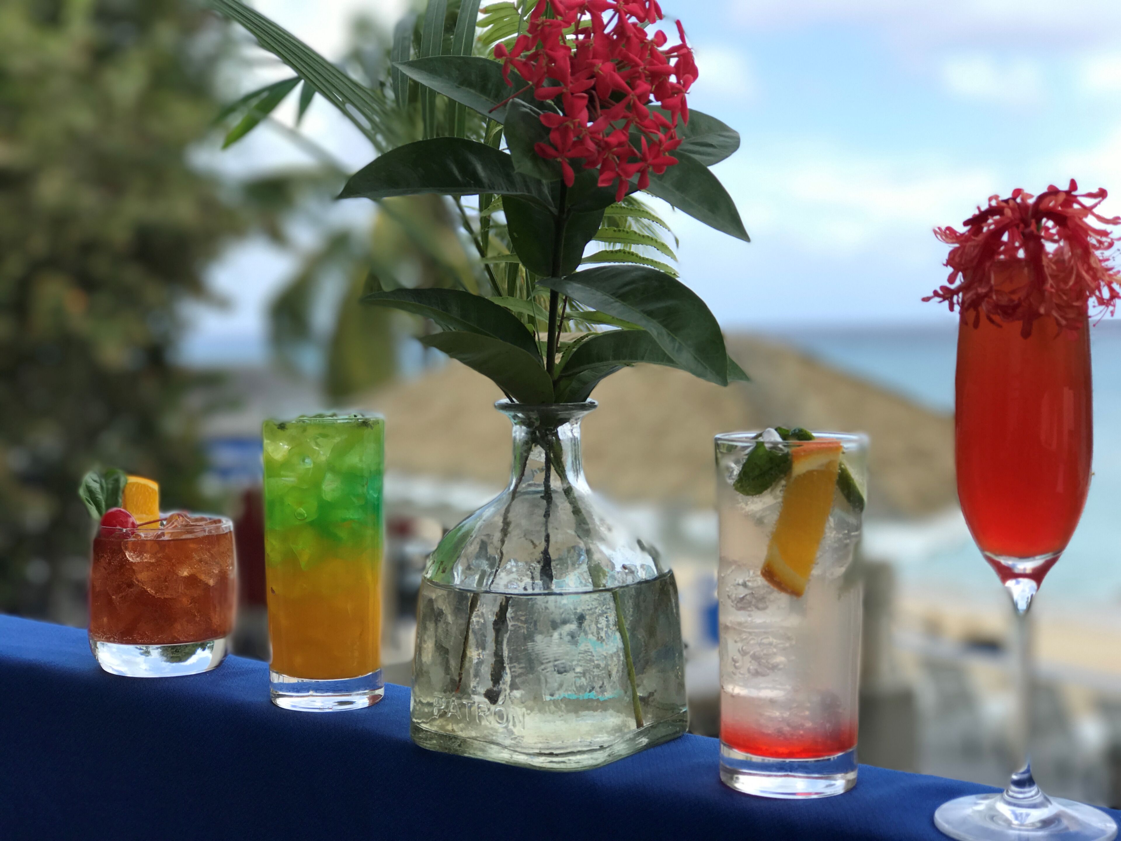 A table with four drinks, a vase with red flowers, and a view of the ocean.