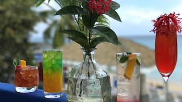 A table with four drinks, a vase with red flowers, and a view of the ocean.