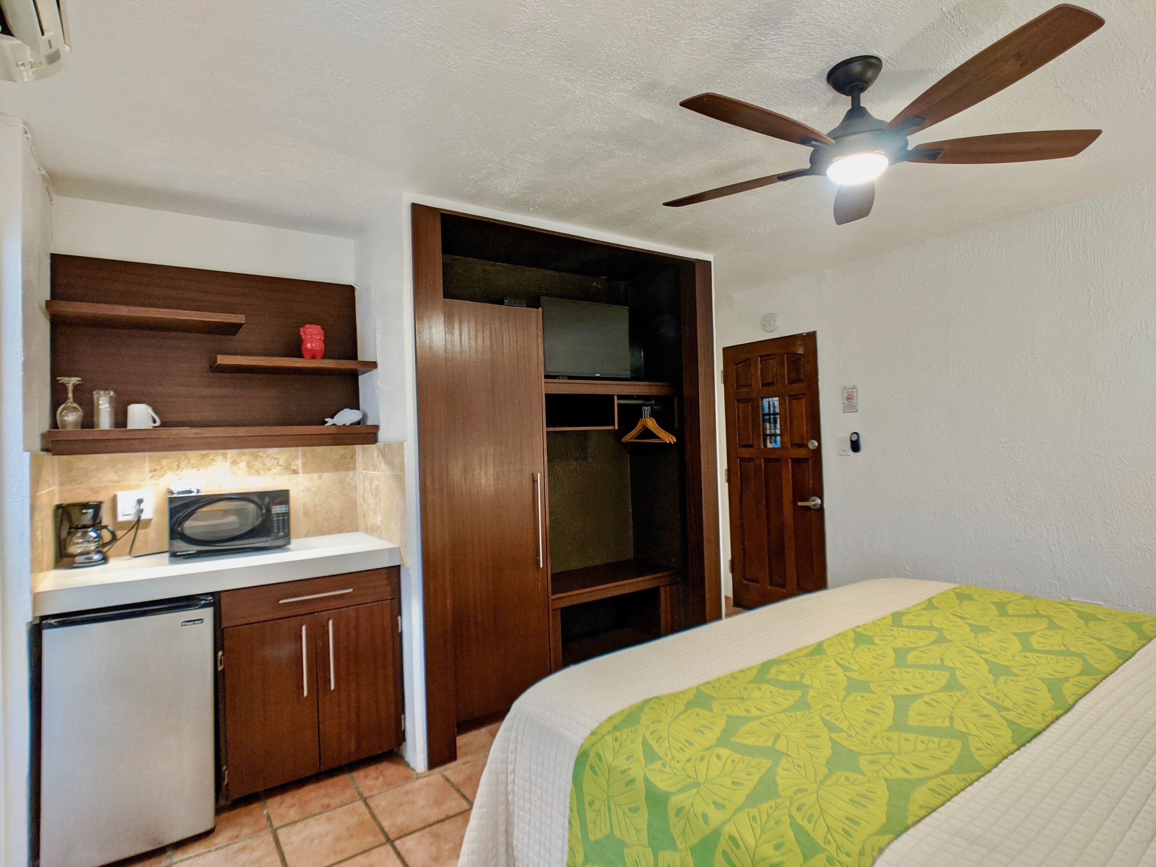 A compact room with a bed, a small kitchenette, a ceiling fan, and a wardrobe.