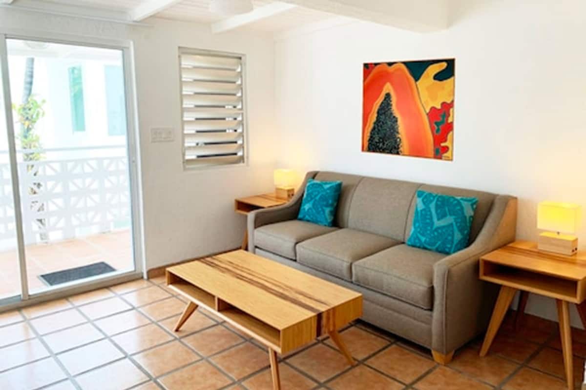 Un salón con un sofá, una mesa de centro de madera y dos mesitas de noche con lámparas. Hay una ventana con persianas y una puerta que da al exterior.