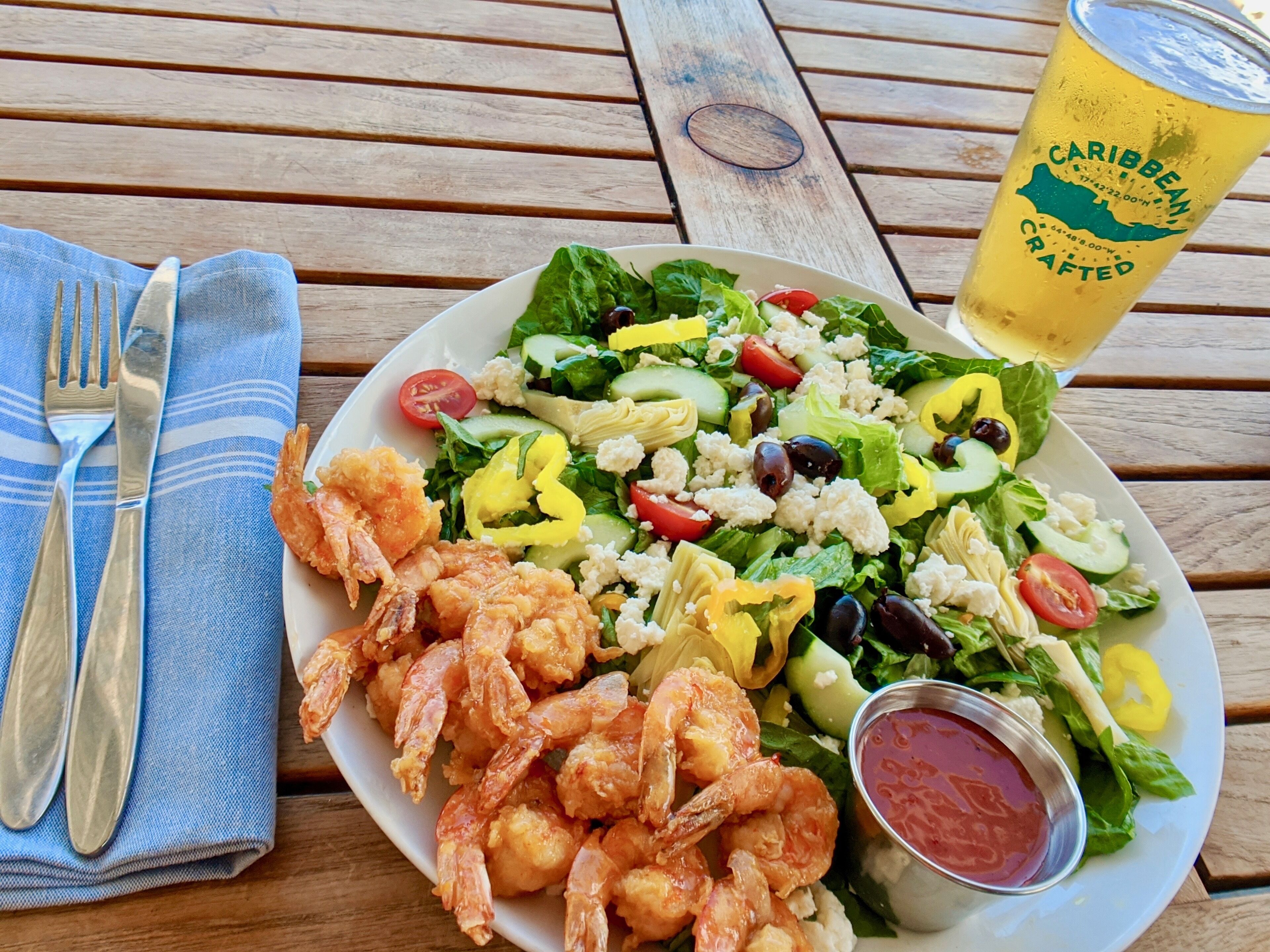 A plate of shrimp and salad with a drink and condiments on a wooden table.
