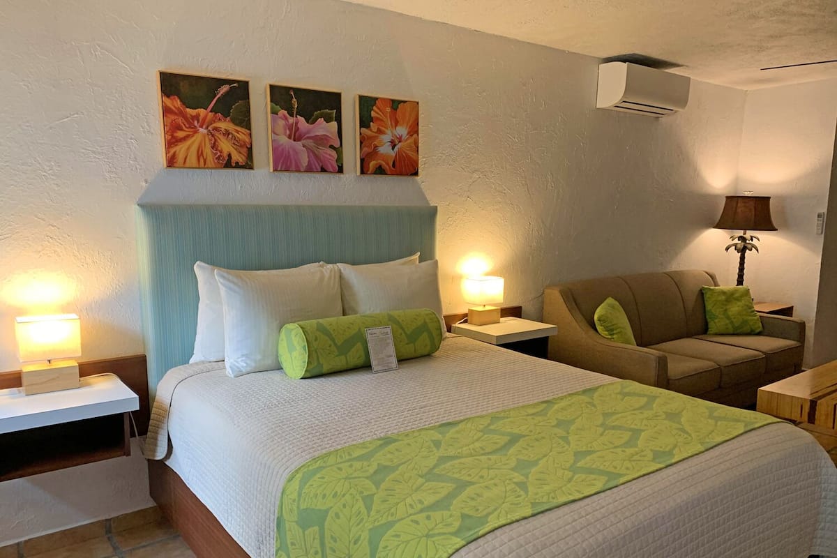 Una habitación de hotel con cama, dos mesitas de noche, un sofá y aire acondicionado instalado en la pared.