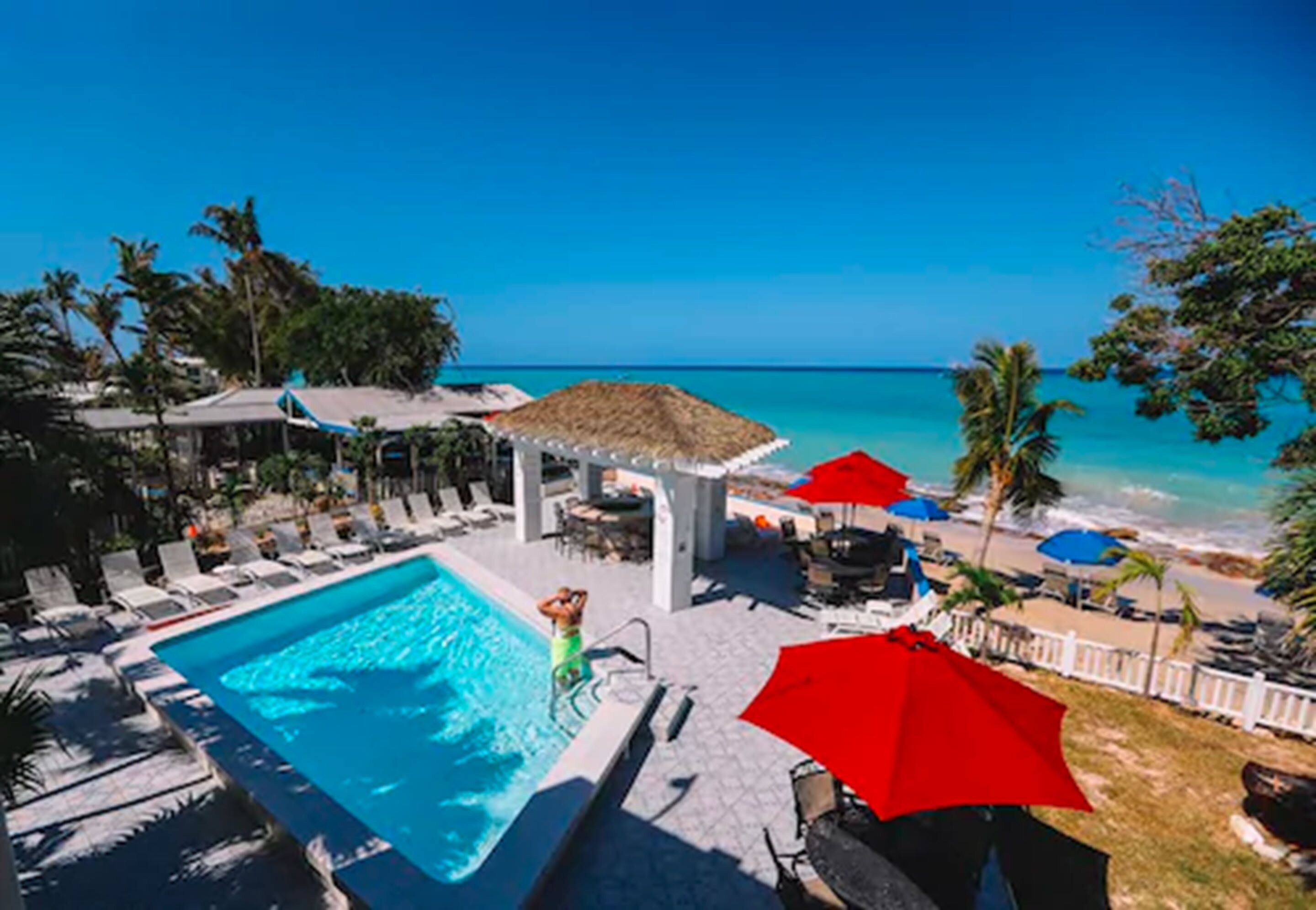 A pool area with lounge chairs and umbrellas, overlooking a beach with palm trees and a clear blue sky.
