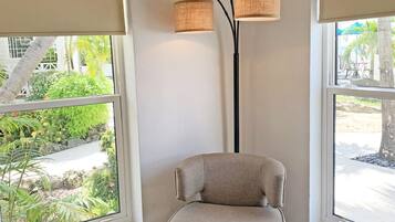 Een kamer met een beige fauteuil, een hoge vloerlamp en grote ramen met uitzicht op een tuin en terras.