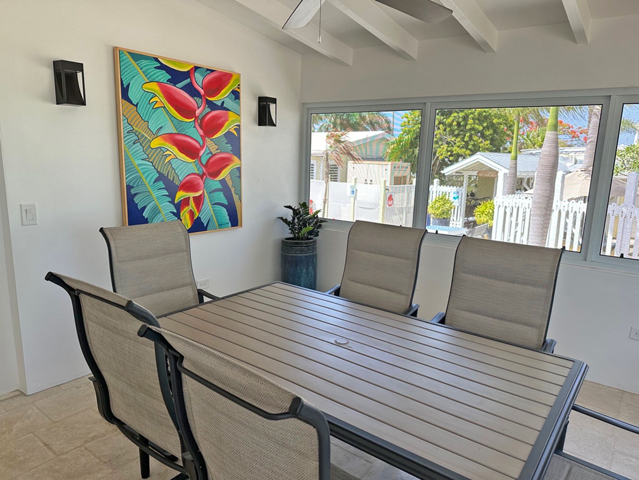A dining area with a table and chairs, a painting on the wall, and a view of the neighborhood through large windows.