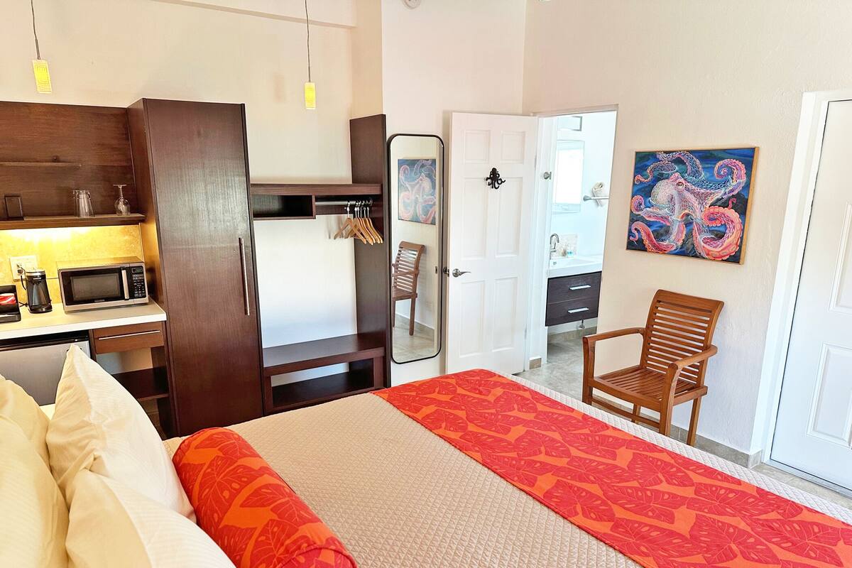 Habitación de hotel con cama, escritorio con microondas, silla, armario y baño visible a través de una puerta abierta.