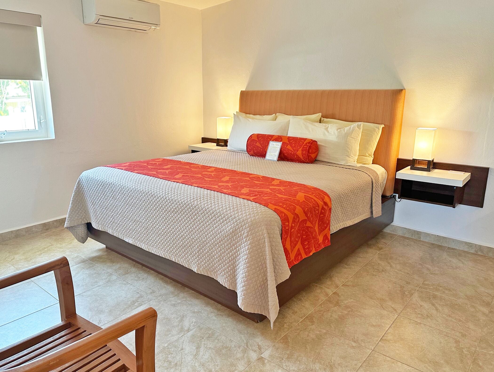 A neatly made bed with a patterned blanket, two bedside tables with lamps, and a wooden chair in a hotel room.
