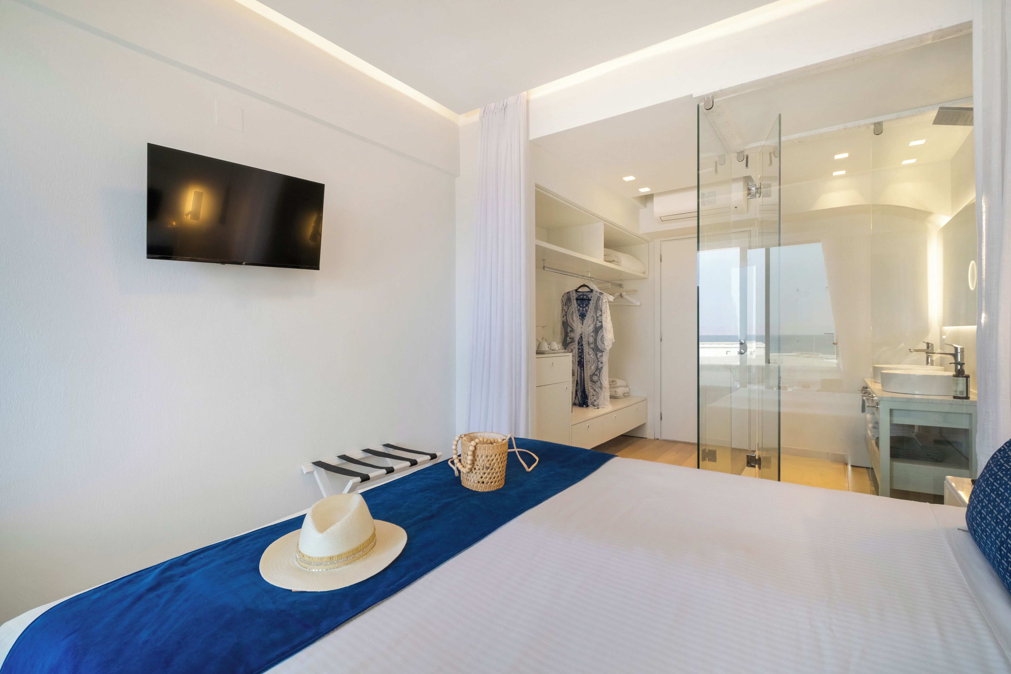 A modern hotel room with a bed, a TV mounted on the wall, a walk-in closet, and a bathroom with a glass shower.
