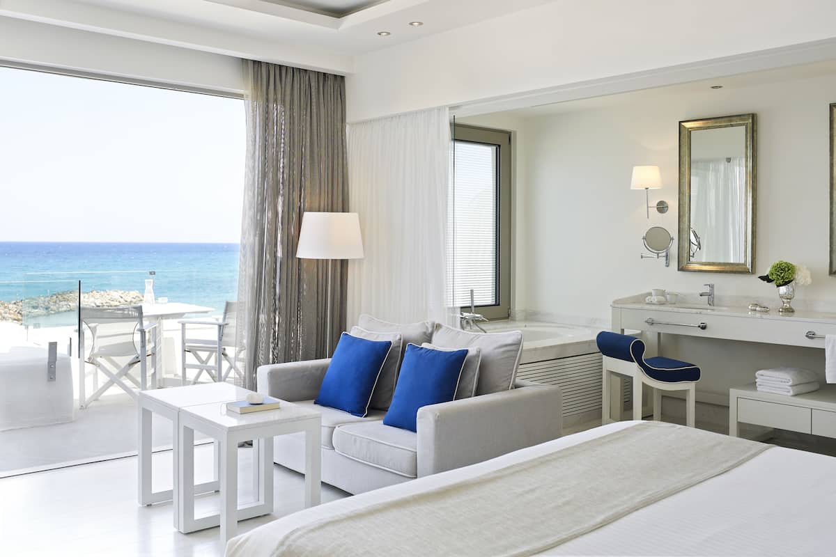 Ein Hotelzimmer mit Bett, einer Couch mit blauen Kissen, einem kleinen Tisch, einem Schreibtisch, einem Badezimmer mit Spiegel und Waschbecken sowie Blick aufs Meer.