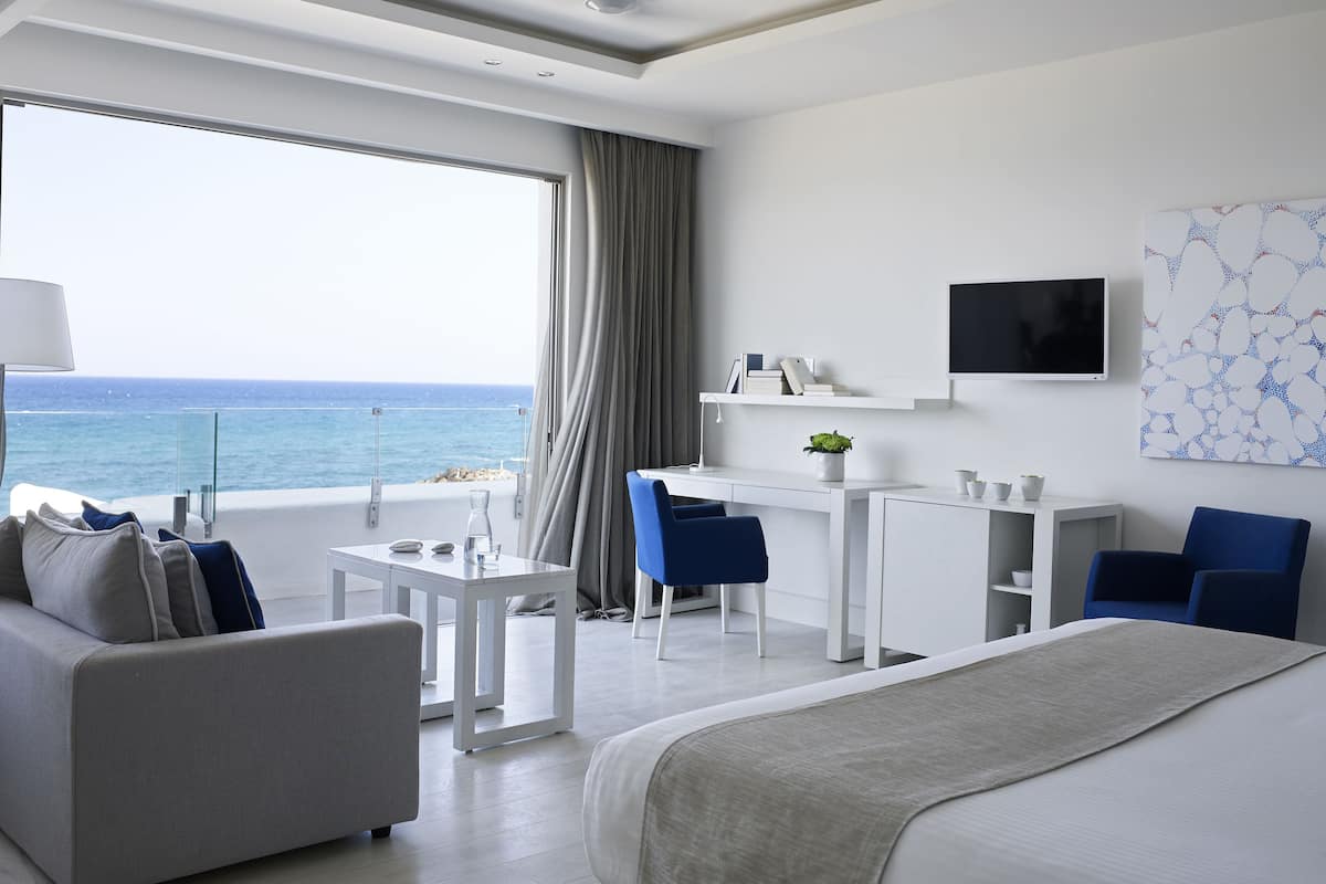 Ein modernes Hotelzimmer mit einem großen Bett, einer Couch, einem Schreibtisch und einem Sessel. An der Wand befindet sich ein Fernseher und ein Fenster mit Blick auf das Meer.