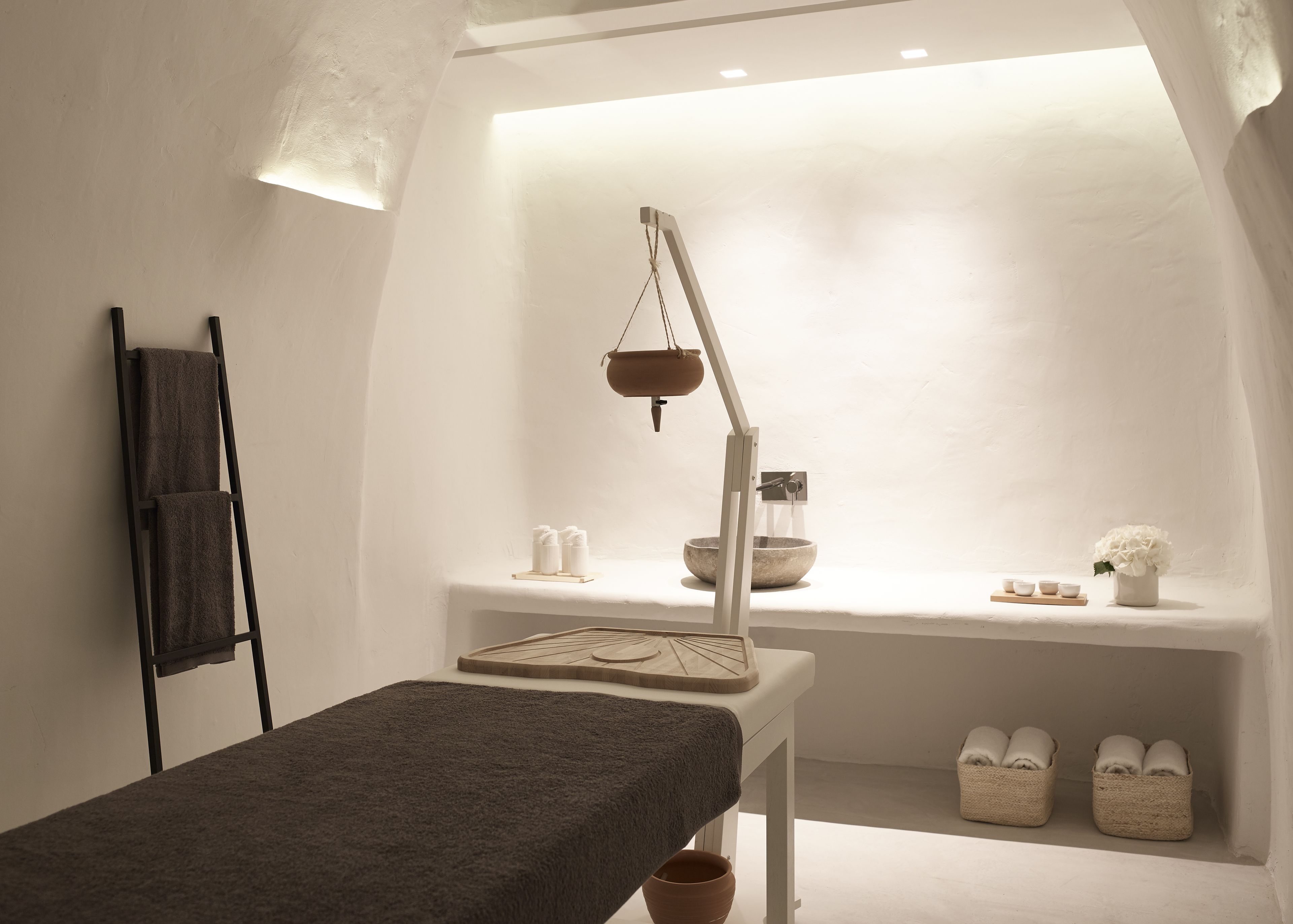 Un cuarto de spa con una camilla de masaje, un estante con toallas y artículos de aseo, y una maceta colgante.