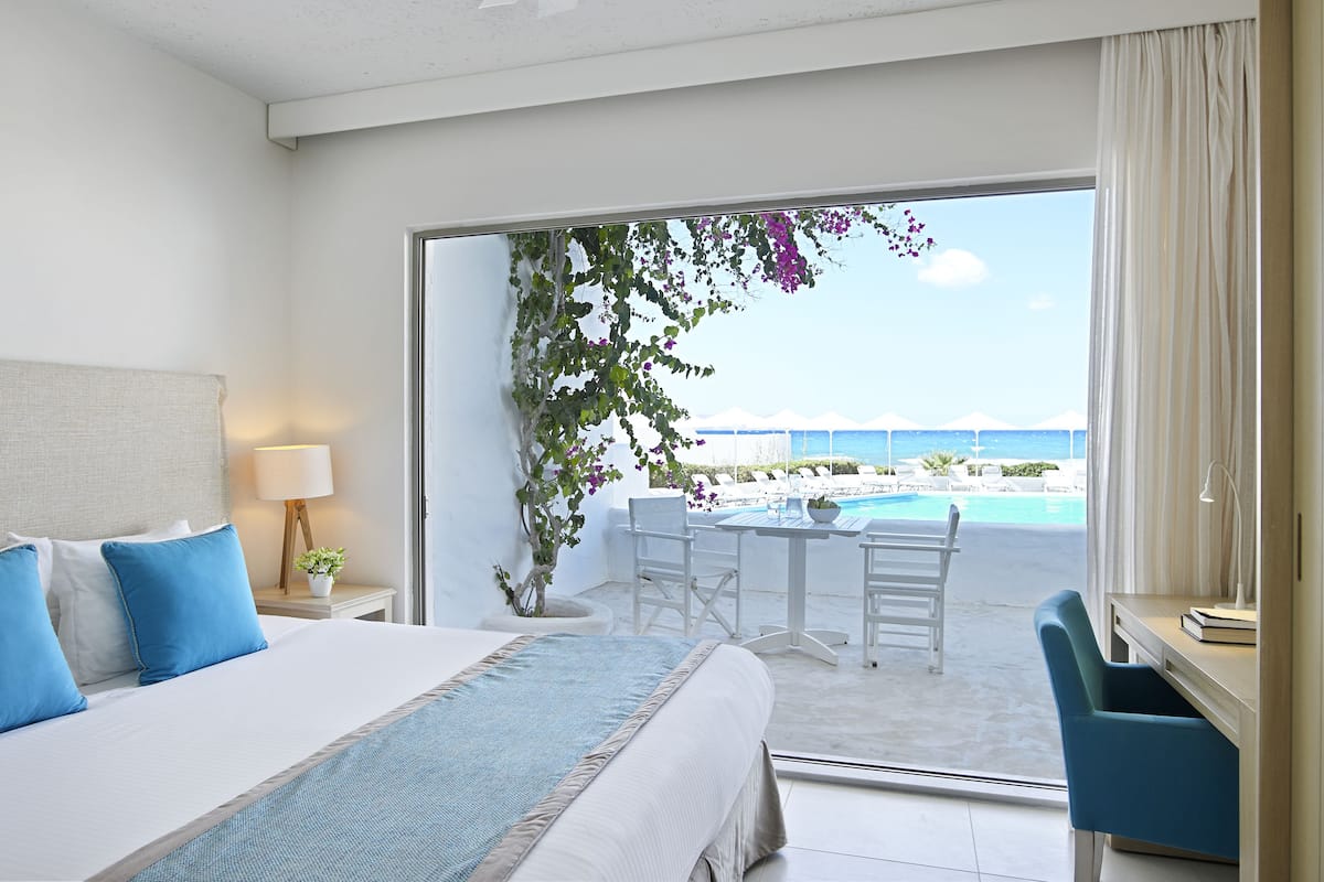 Ein Hotelzimmer mit Bett, Schreibtisch, Stuhl, Lampe, Zimmerpflanze und Blick auf Pool und Strand.