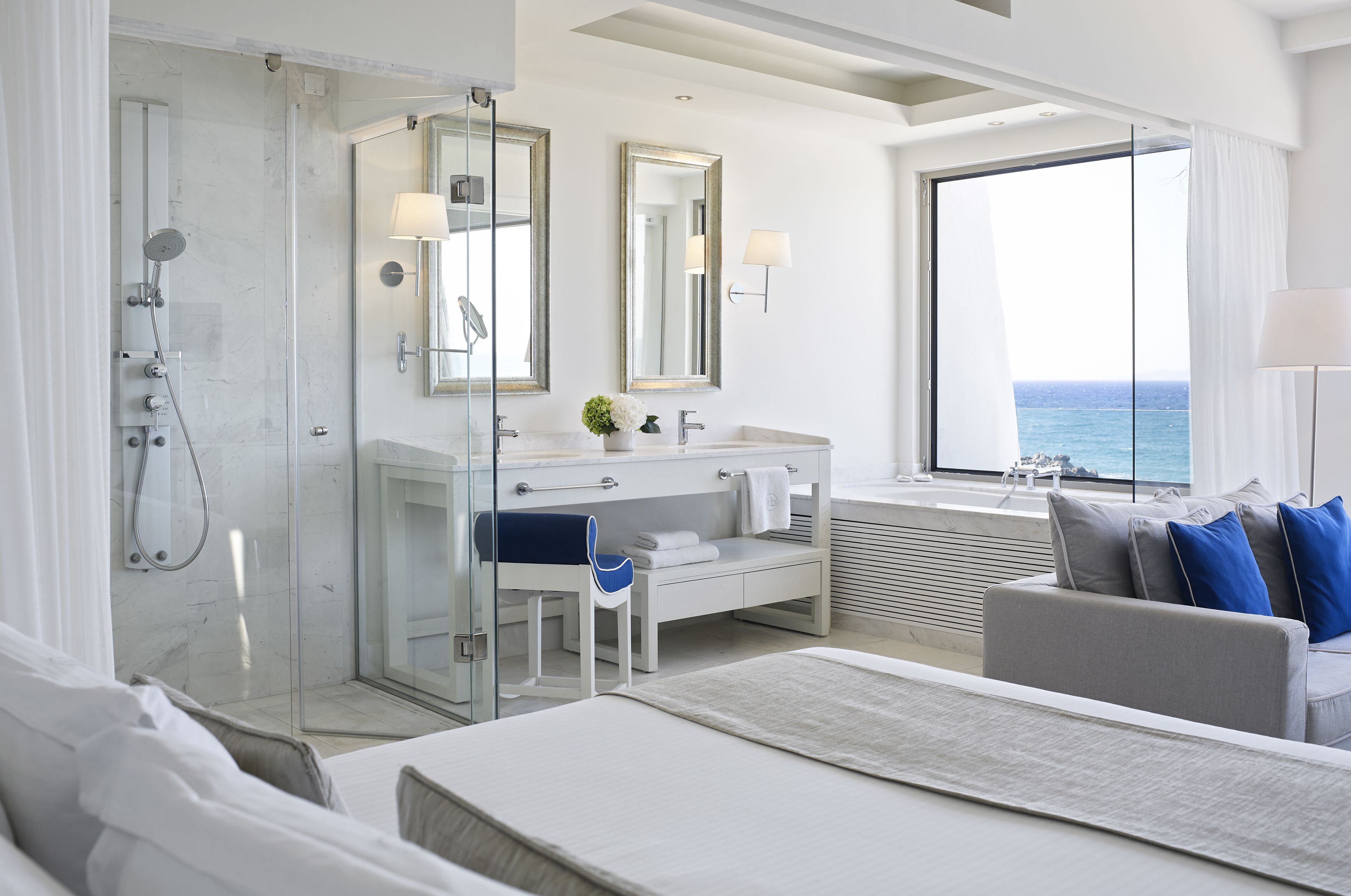 A modern hotel room with a glass-enclosed shower, a large window with a view of the sea, a white vanity with a mirror, and a bed with a view of the ocean.