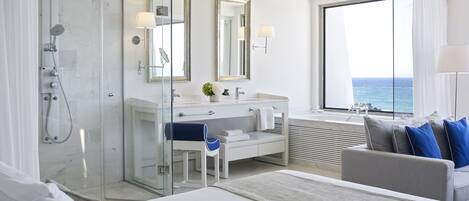 A modern hotel room with a glass-enclosed shower, a large window with a view of the sea, a white vanity with a mirror, and a bed with a view of the ocean.