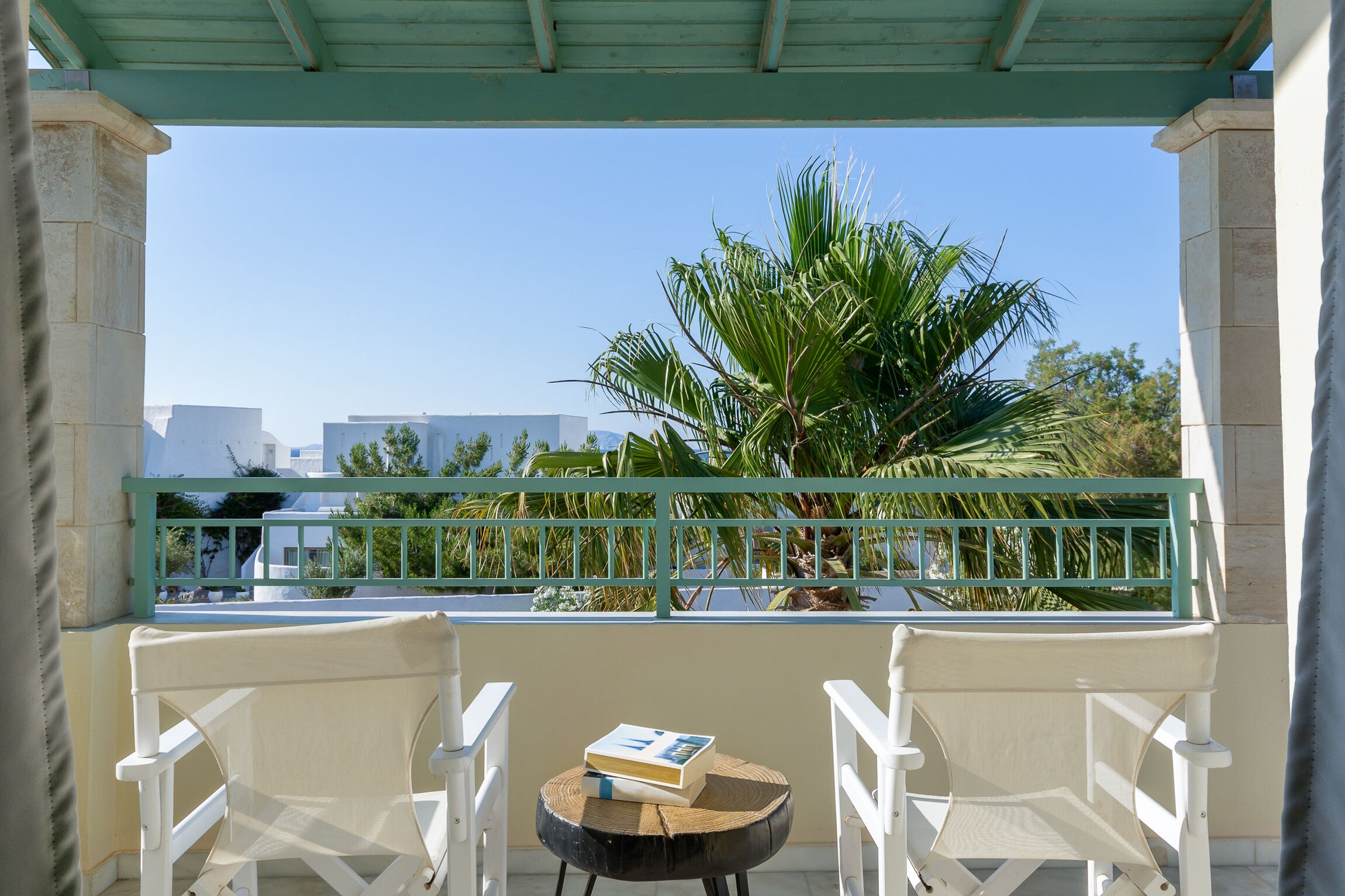 A balcony with white chairs and a small round table, a palm tree, and a view of buildings in the background.