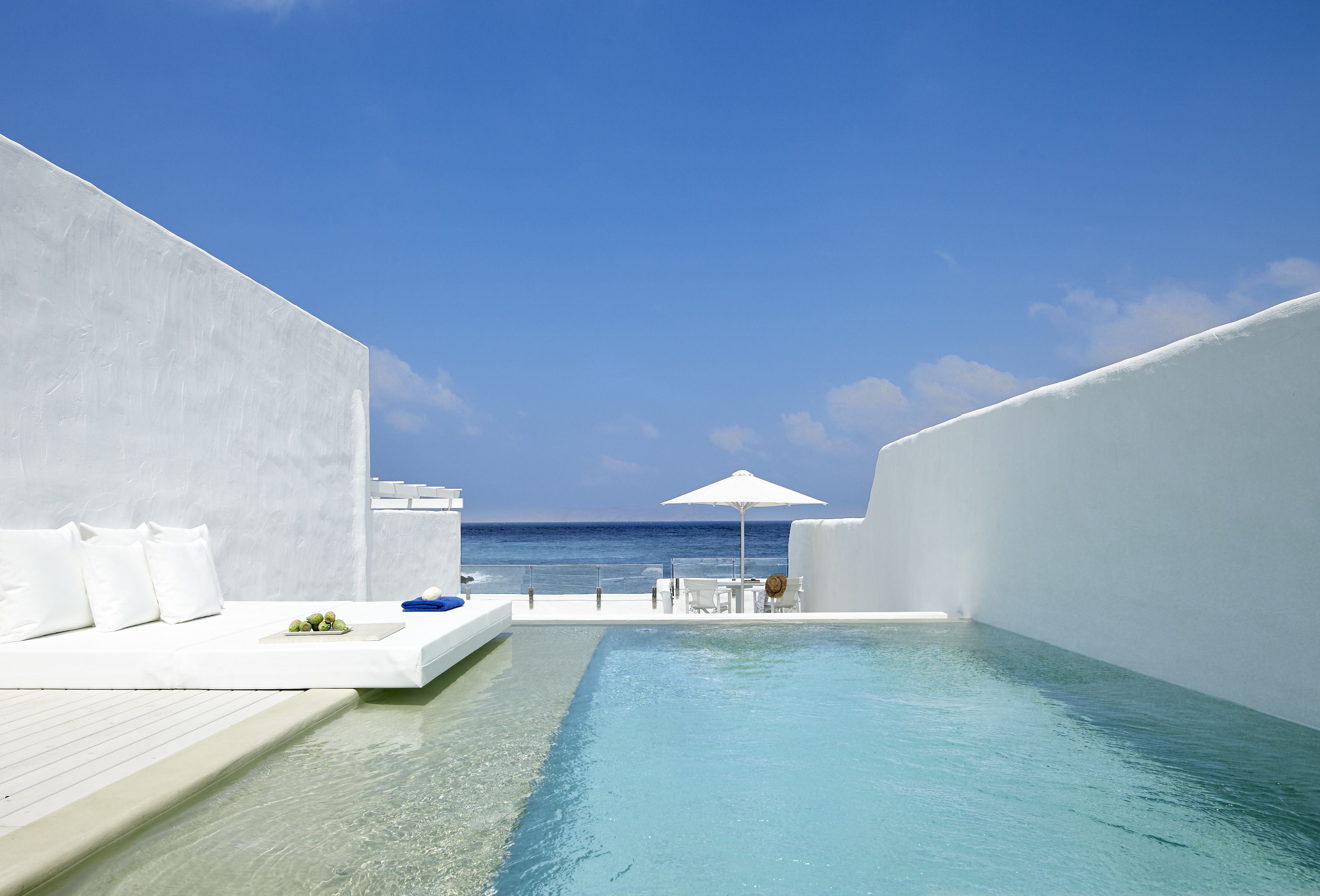 Una piscina infinita moderna con vista despejada al océano, paredes blancas y un área de estar exterior minimalista.