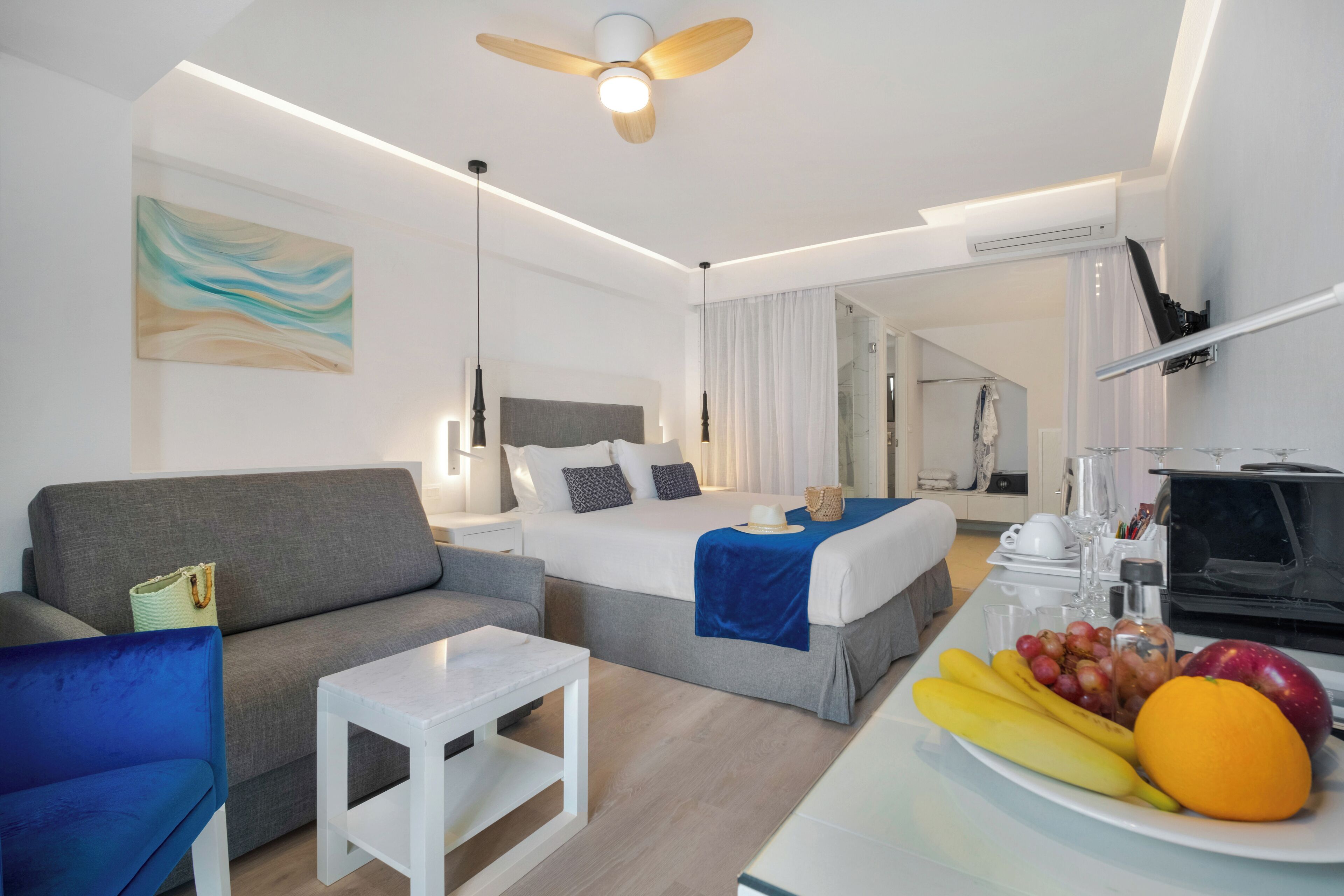 A modern hotel room with a bed, sofa, and a dining table with fruit.