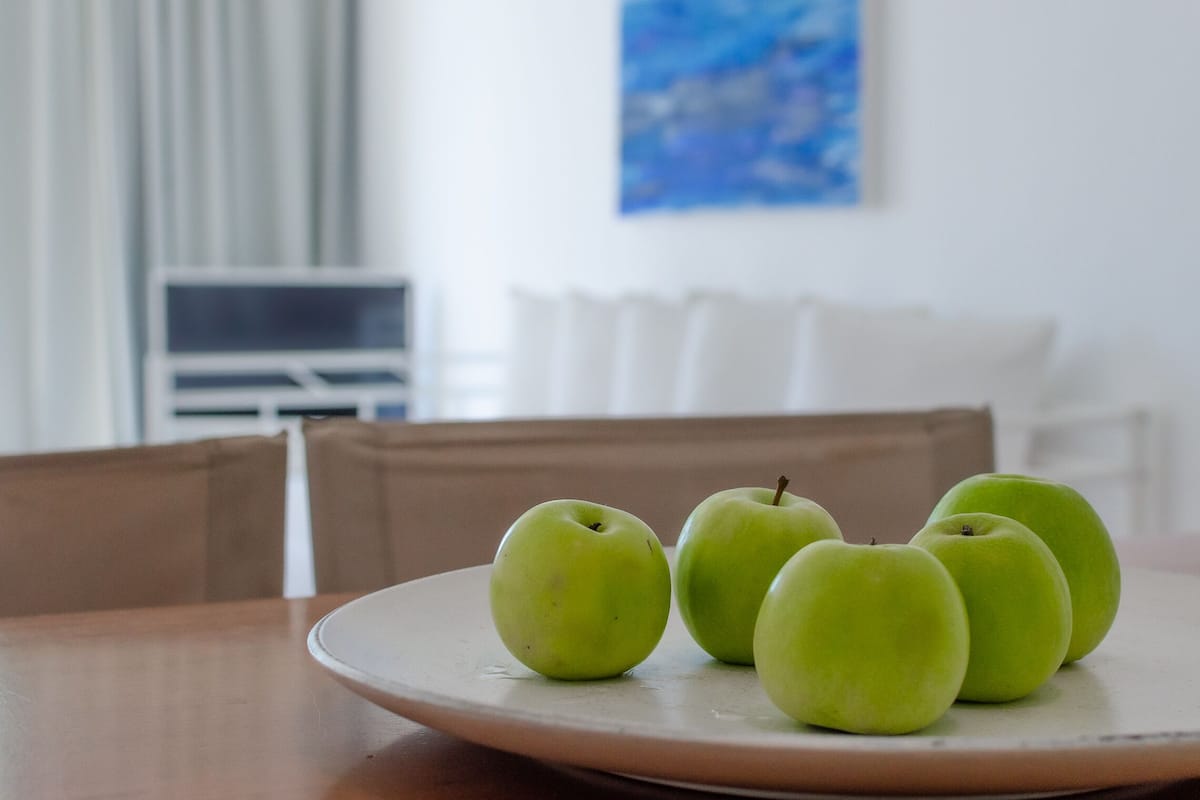 Ein Teller mit vier grünen Äpfeln auf einem Holztisch in einem Raum, der ein weißes Sofa und ein Bild an der Wand zeigt.