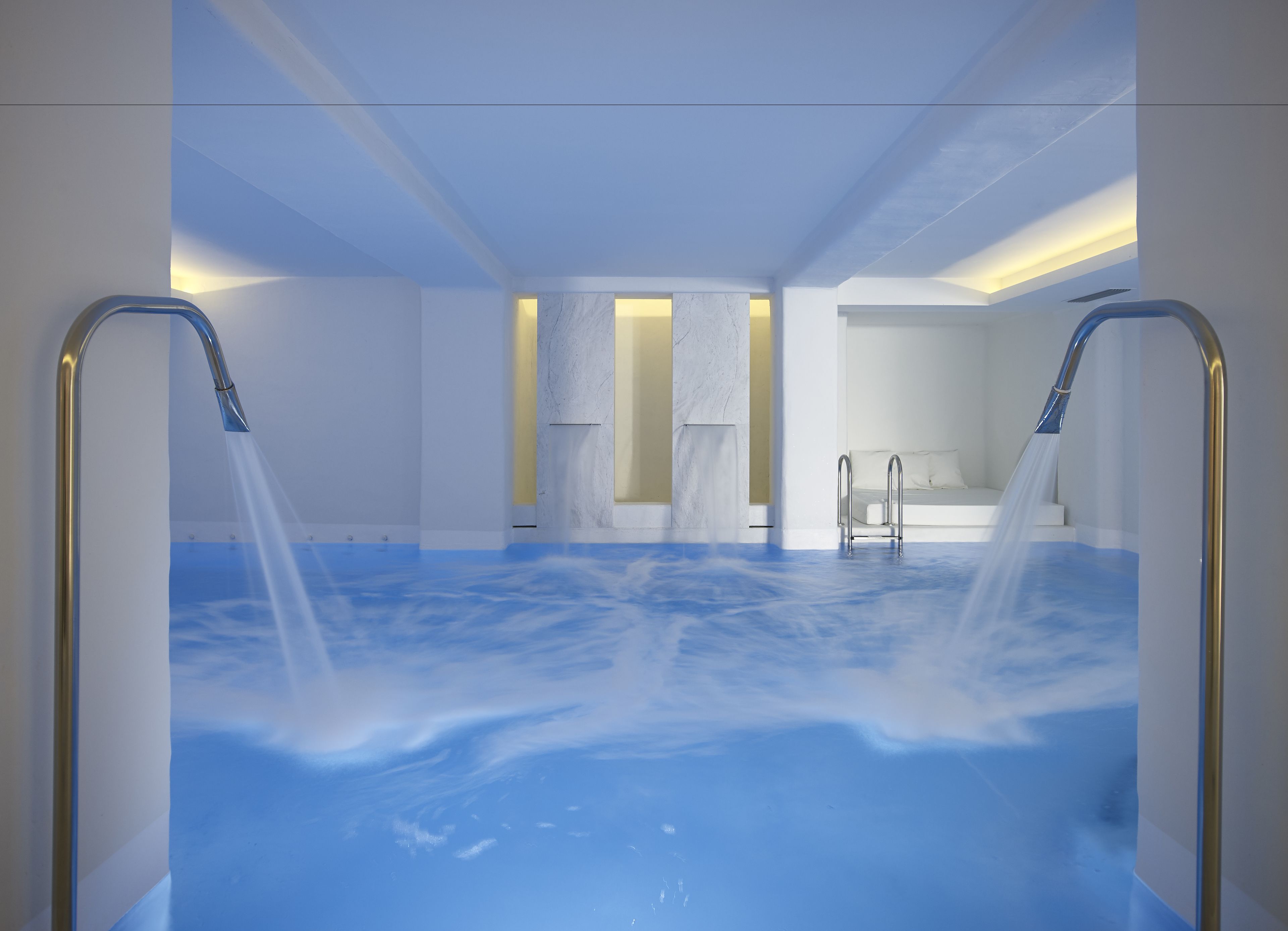 A modern spa room with a large blue heated pool, two showerheads, and a bed with a view of the sky.