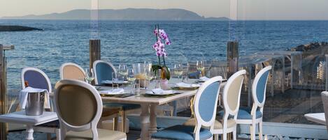 Comedor con una mesa dispuesta para seis personas, con vista a una masa de agua y un litoral rocoso.