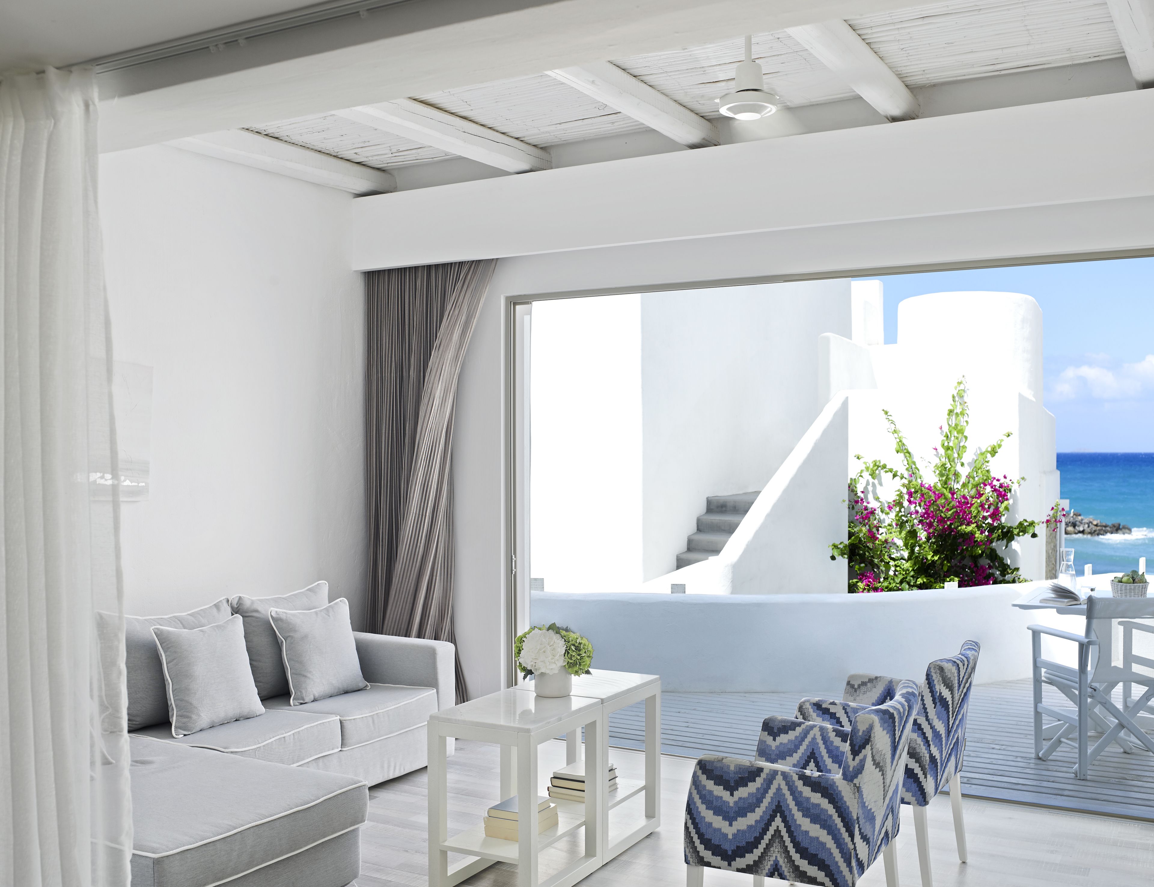 A modern living room with a grey sofa, a white coffee table, and patterned chairs. A staircase leads to a balcony with a view of the ocean.