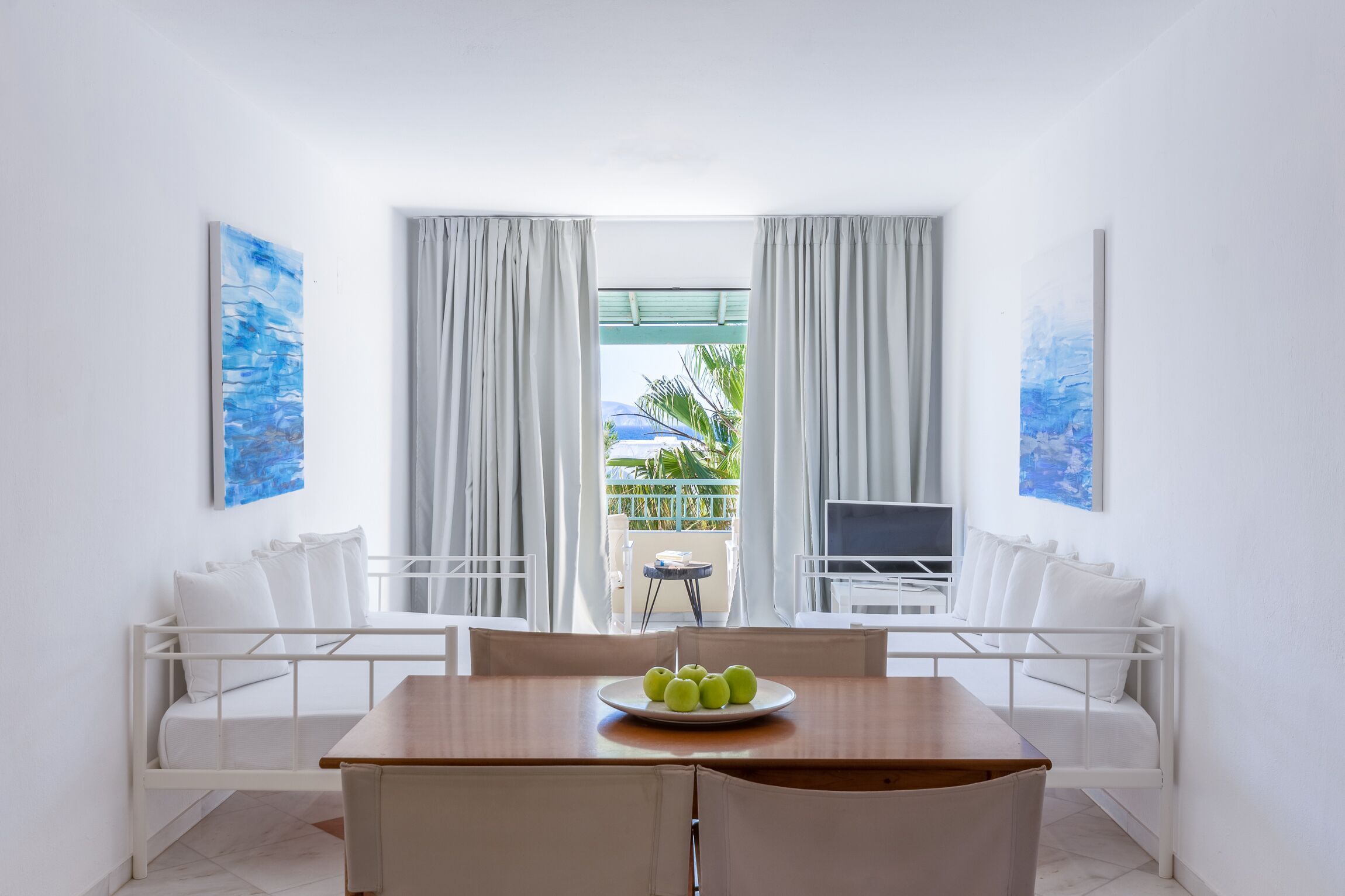 A room with a dining table, white chairs, a white bed, and a balcony with a view of palm trees.