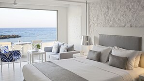 A modern hotel room with a large bed, a balcony with a view of the sea, and a dining area with a table and chairs.